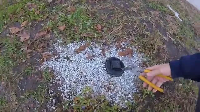 Black cleanout cap in a bed of white gravel with a hand holding a yellow pencil next to it.