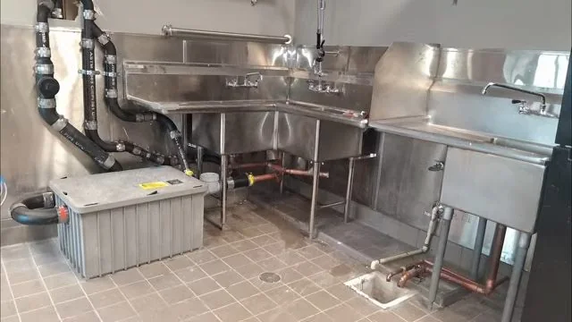 Commercial kitchen corner with stainless steel sinks, black drain pipes and a grease interceptor box on a tile floor.