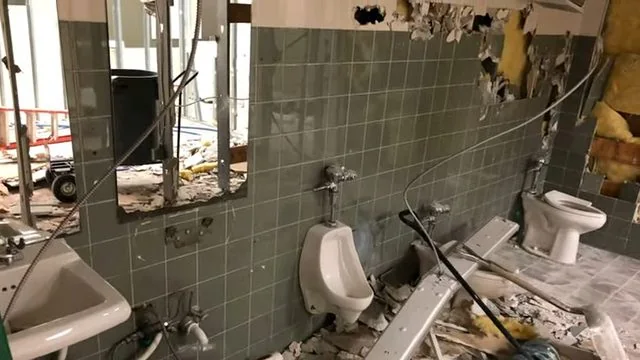 Gutted commercial restroom with tiled wall, urinals, a toilet and exposed pipes.