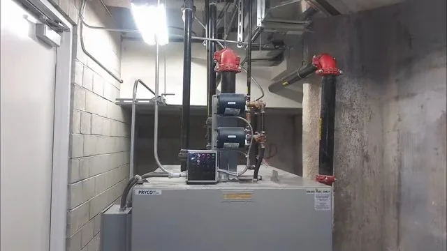 Large commercial water heater with red-handled valves and black pipe in a block-walled utility room.