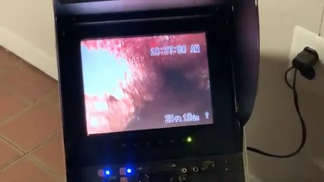 Handheld camera monitor displaying the inside of a drain pipe, resting on a tile floor.