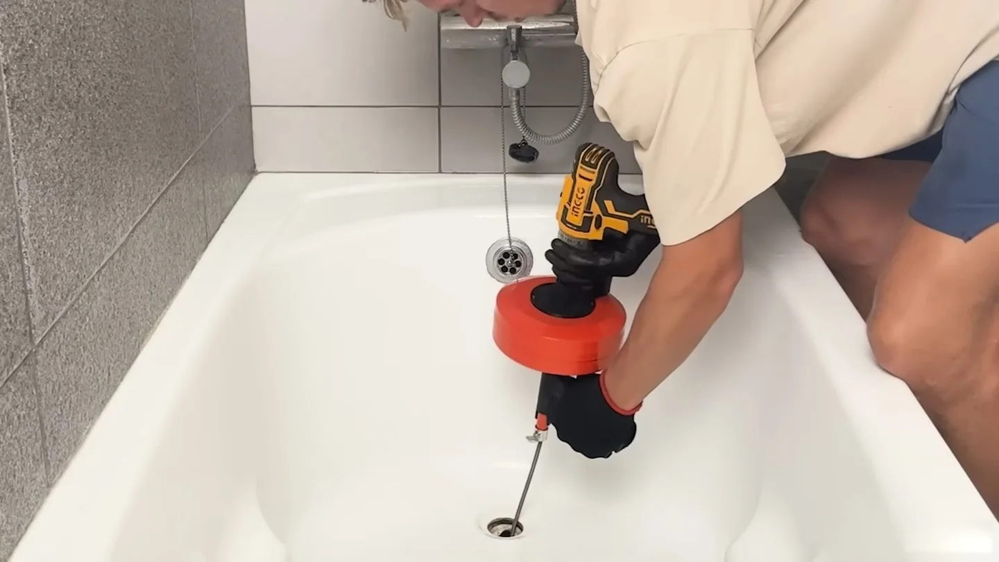 Person using a hand-held drain snake with an orange housing in a white bathtub drain.