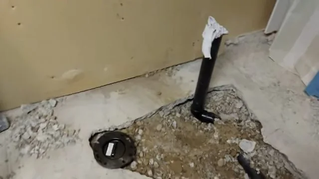 Black drain stub and a round fitting in a rough opening cut into a concrete floor, with rubble around it.