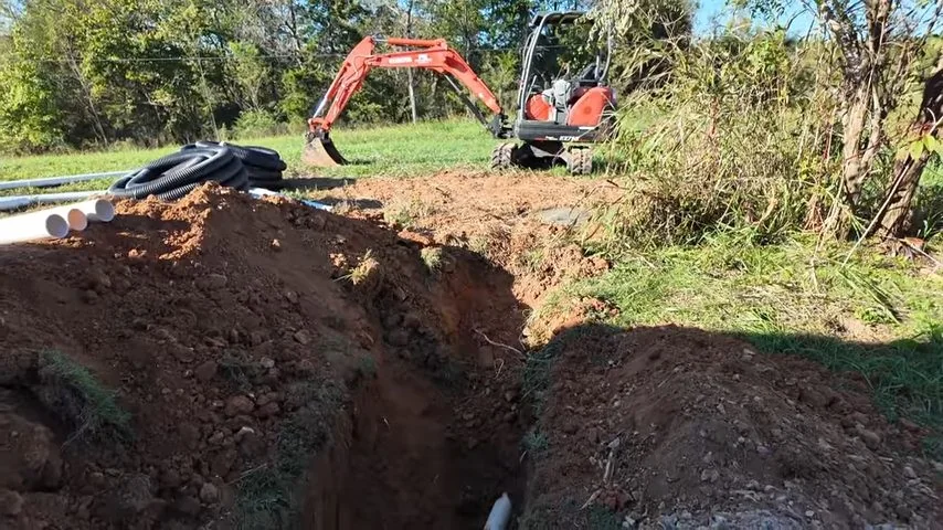 Orange excavator beside a long open trench in a grassy field.