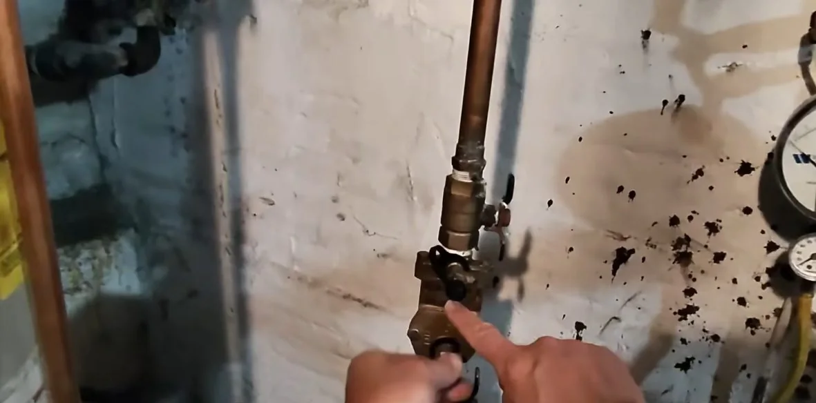 Finger touching a brass valve on a pipe beside a meter and a gauge on a stained wall.