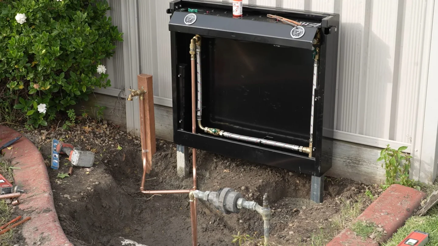 Gas meter and water piping beside a stucco wall with a plant bed and white gravel.