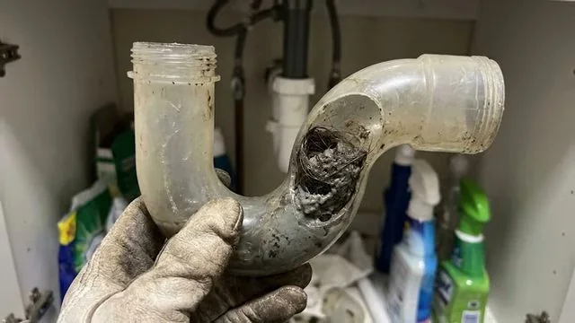 Gloved hand holding a clear P-trap with a crack and grime inside, beside cleaning bottles under a sink.