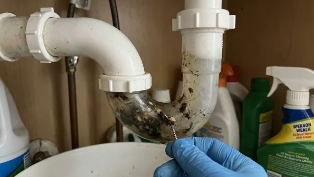 Gloved hand holding a white P-trap with sludge and debris inside, beside cleaning bottles.