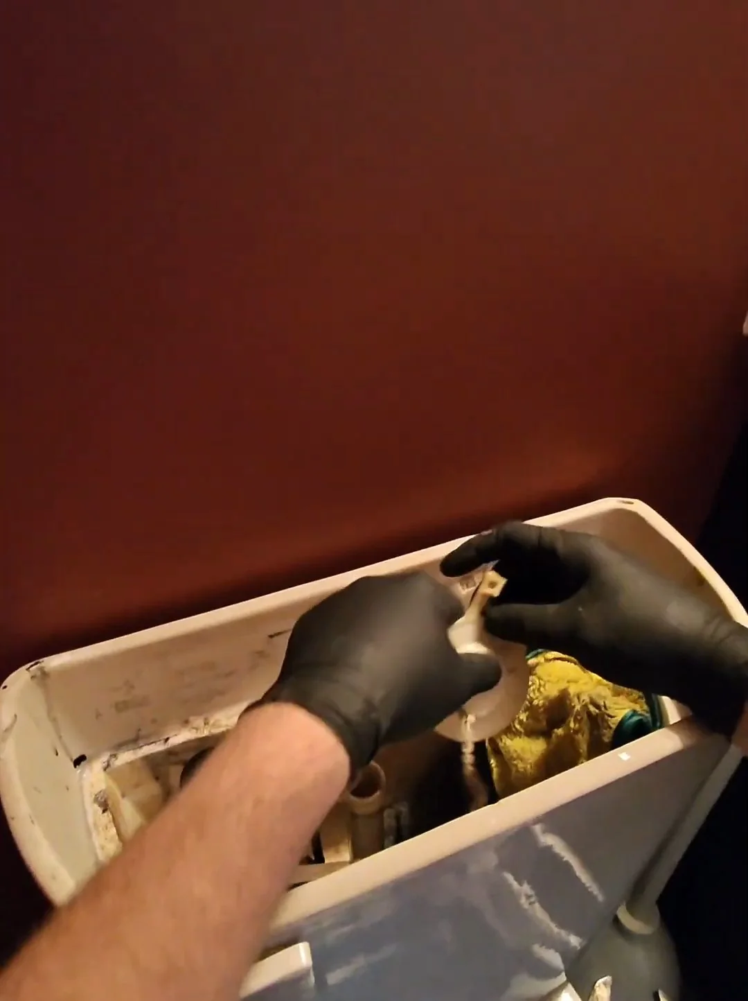 Black-gloved hands working among the parts inside an open toilet tank.