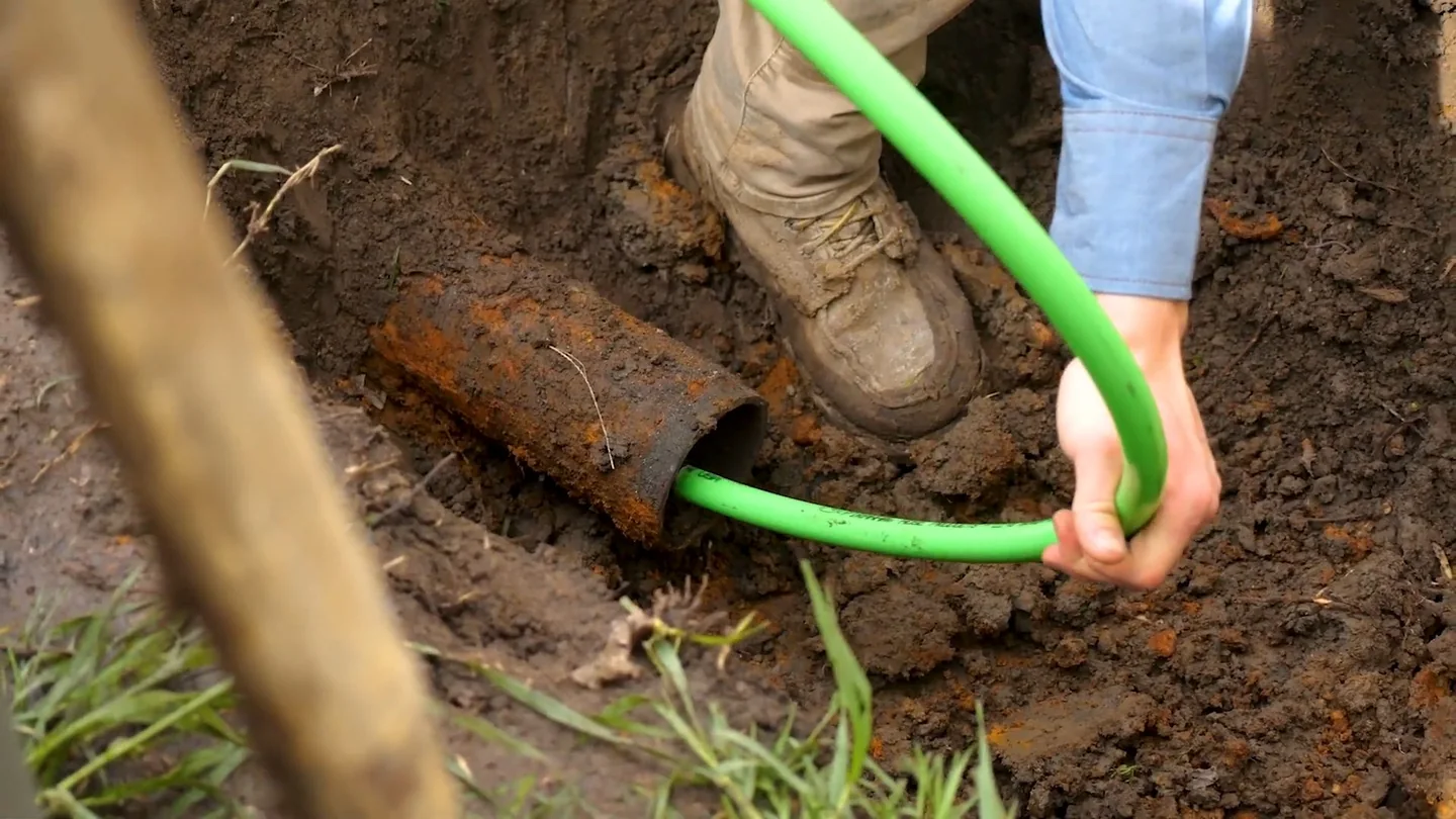 Hand feeding a green hose into the open end of a buried pipe in dark soil.