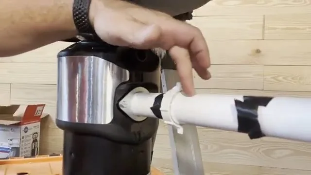Hand holding a white drain pipe against the discharge port of a stainless garbage disposal.