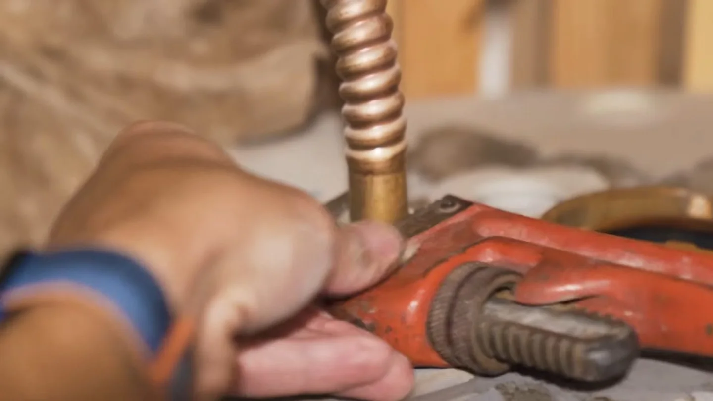 Hand gripping a red pipe wrench on a copper supply line with a corrugated connector above it.