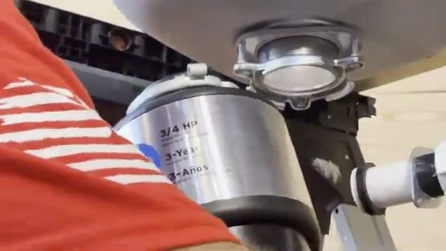 Hand on the side of a garbage disposal unit mounted under a sink, with a white drain pipe leaving it.