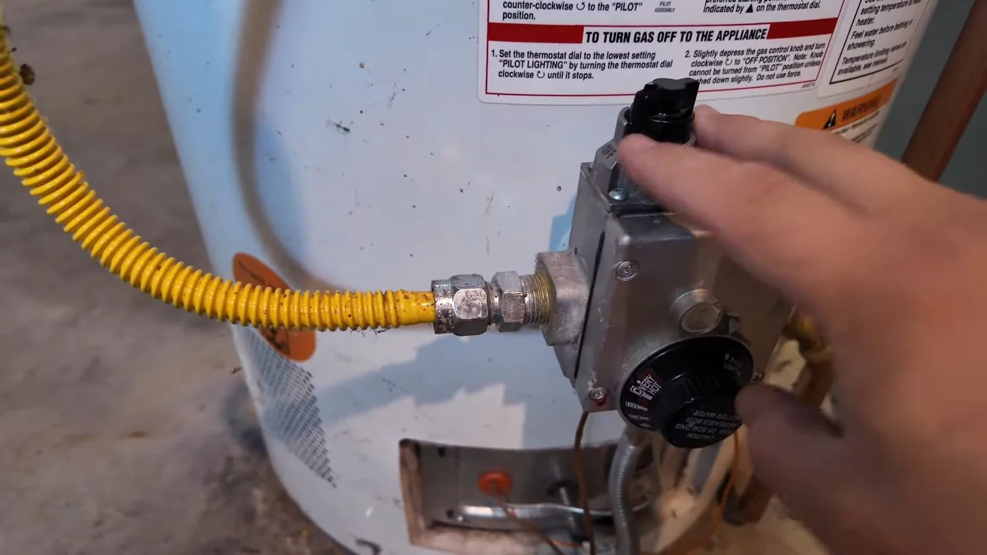 Hand turning a gas control on a water heater with a yellow flexible gas line beside it.
