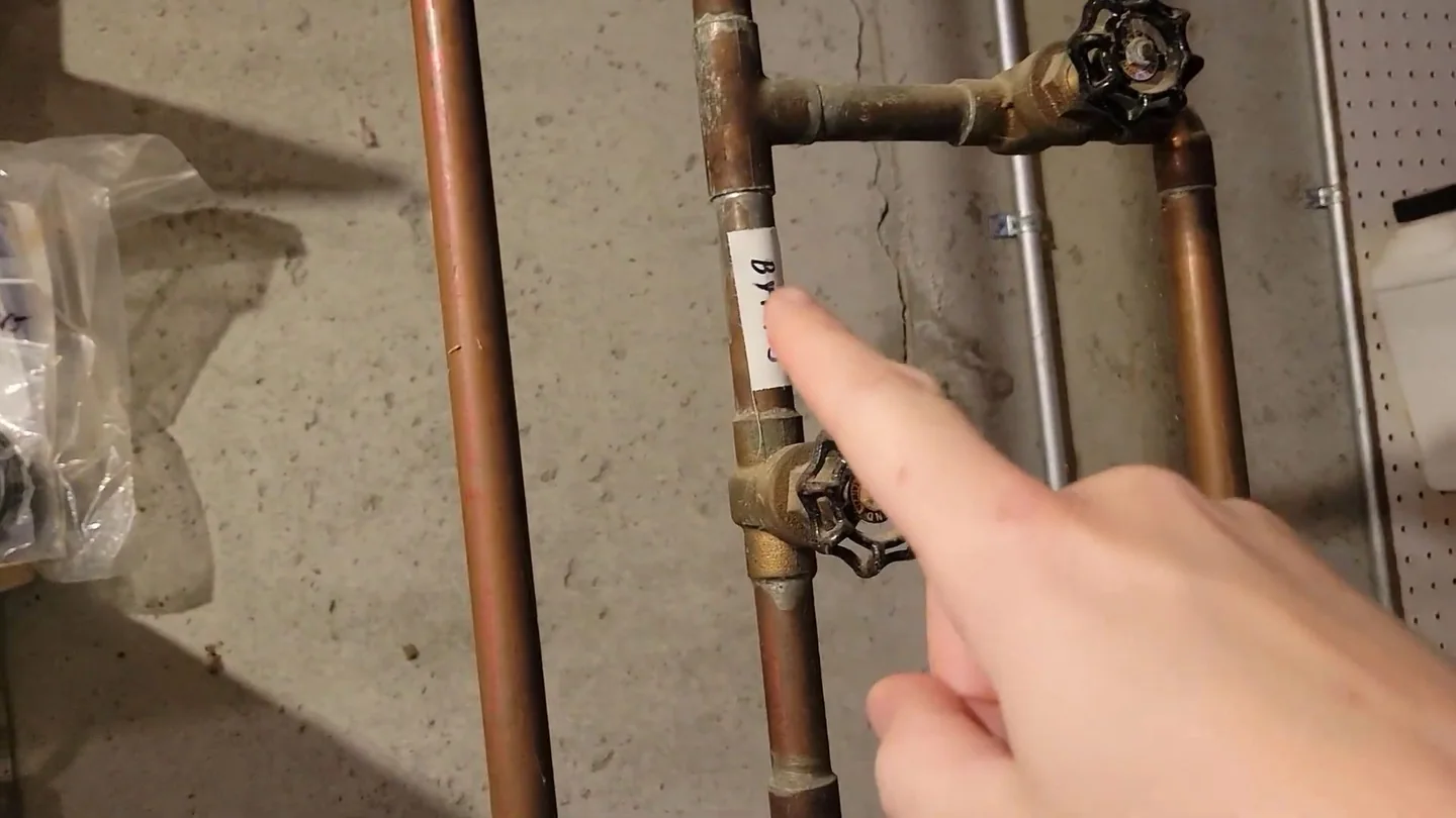 Hand pointing at brass valves on copper supply lines running along a concrete wall.