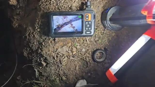 Handheld camera monitor with a live image lying on the soil beside a pipe and a reel.