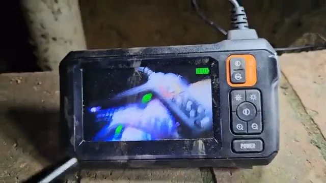 Handheld inspection camera monitor with a pipe view on screen and an orange button panel.