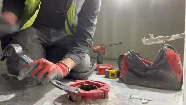 Kneeling worker in gloves with a red wax ring, a wrench and a tool bag on a bare floor.
