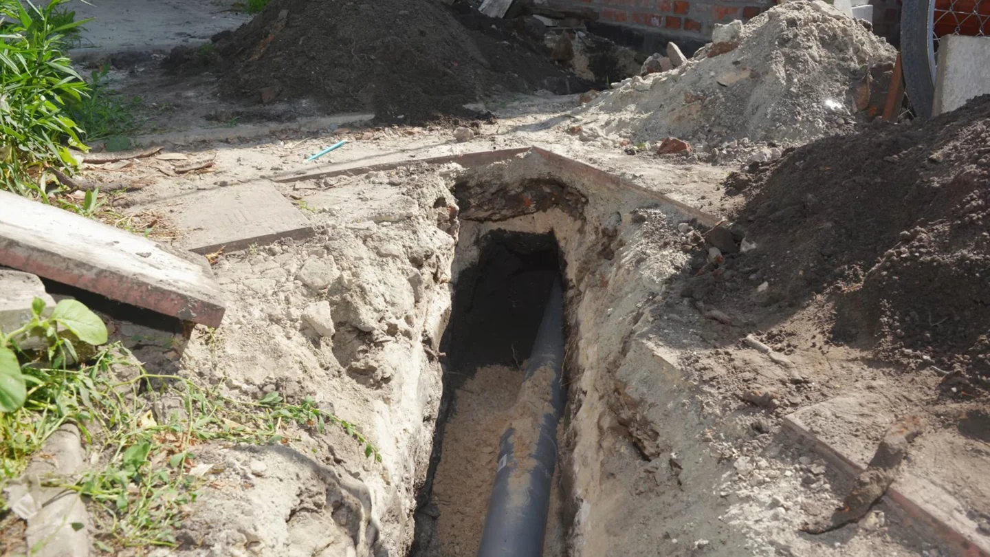 Narrow trench in sandy soil with a dark sewer pipe visible along its bottom and a pile of fill beside it.