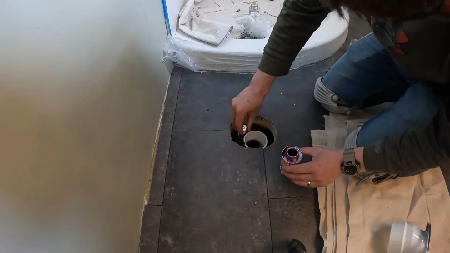 Man crouching beside a toilet at a floor opening, working with a tool near the drain.