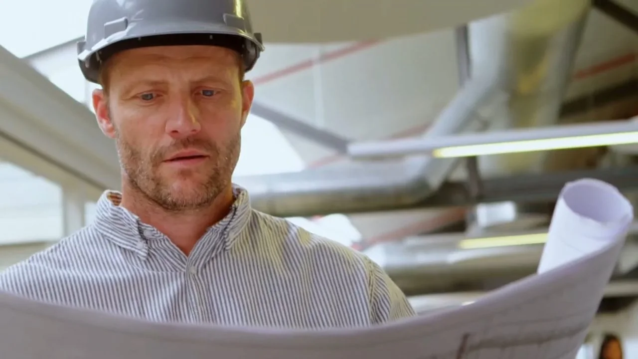 Man in a hard hat studying rolled plans in a space with pipes overhead.