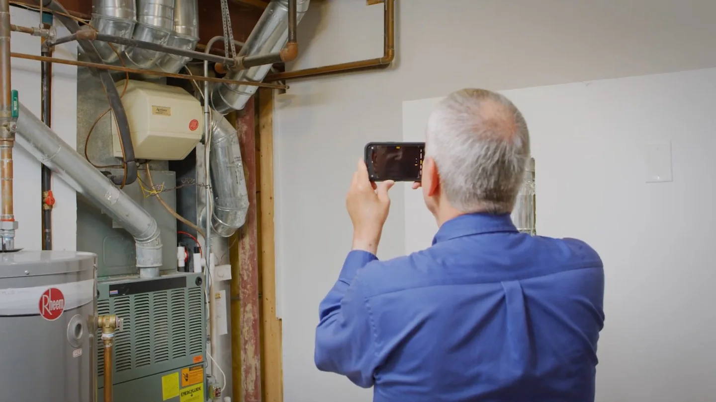 Man in a blue shirt holding up a phone to photograph a water heater and its piping.