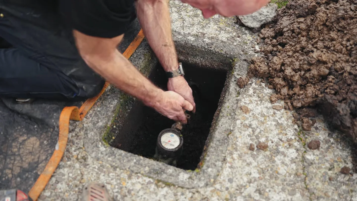 Man leaning over an open ground box to read a water meter with his hand beside the dial.