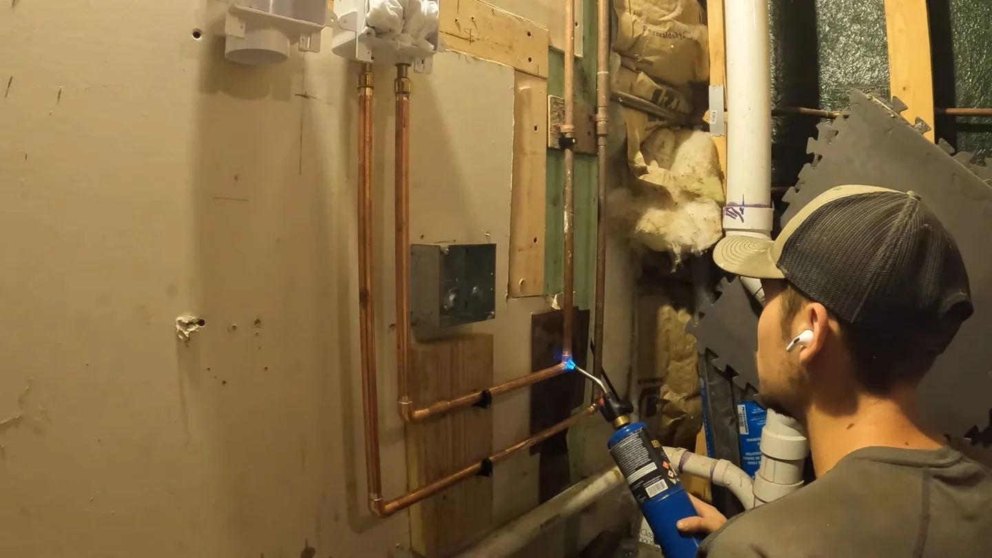 Man in a cap using a torch on copper pipe in a utility space beside a washer.