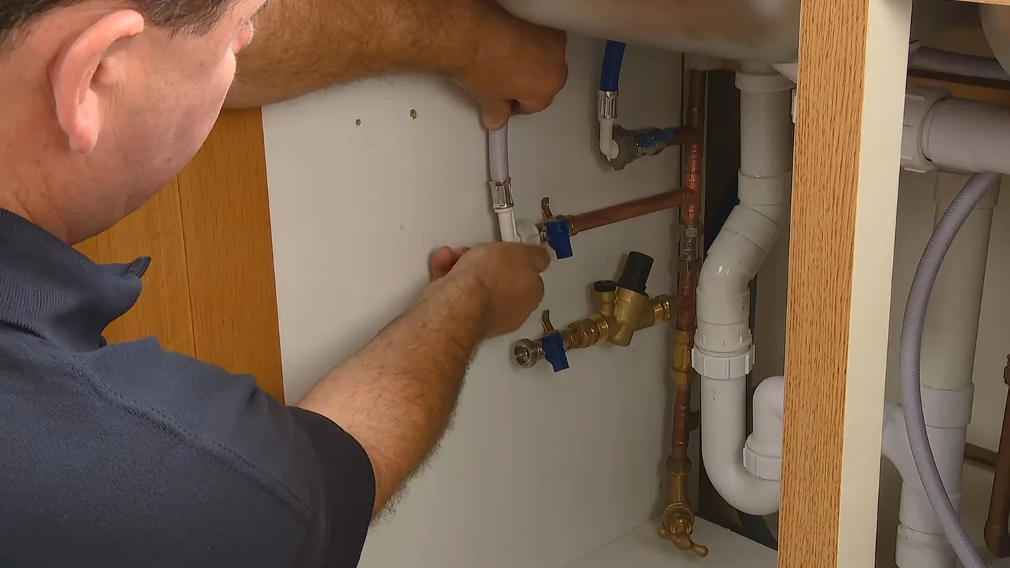 Man in a dark work shirt reaching into a sink cabinet to work at shutoff valves and drain pipes.