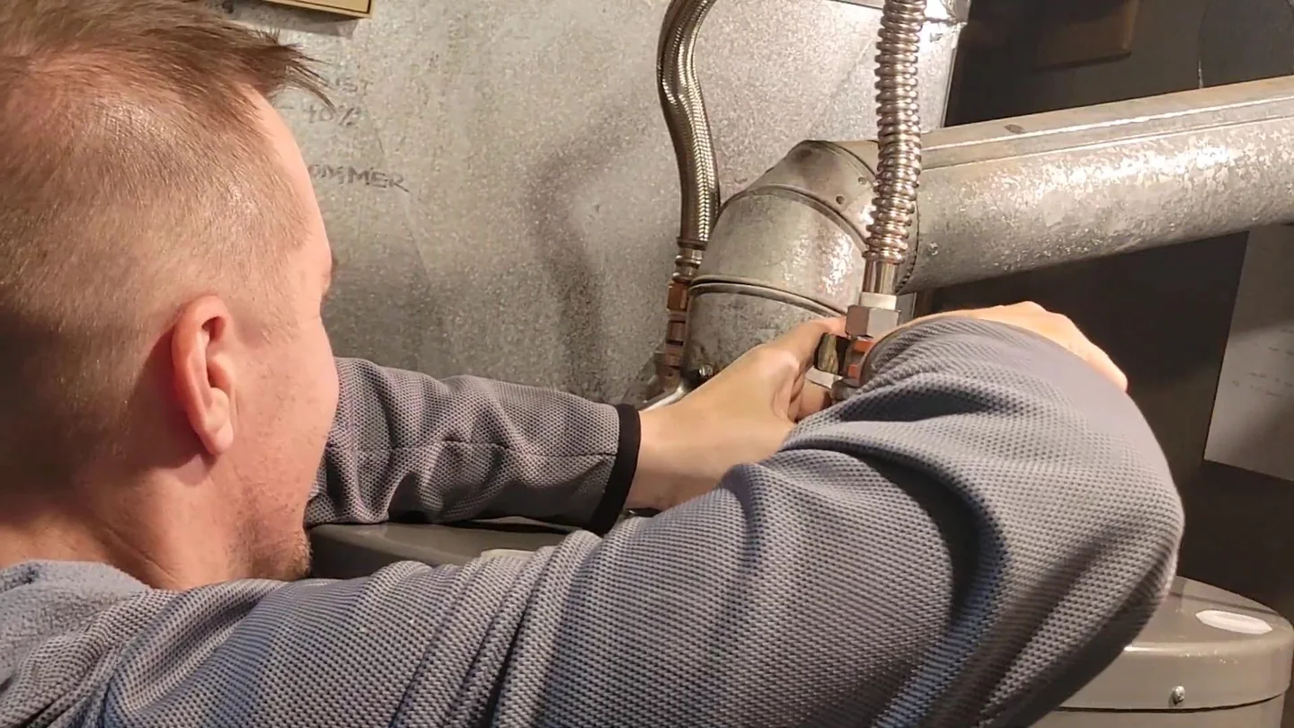 Man with short hair reaching up to metal pipes beside a water heater.