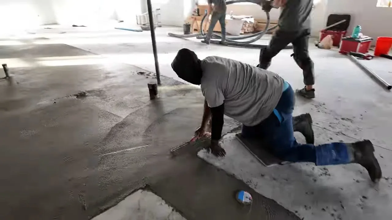 Two men kneeling on a bare concrete floor in a large commercial space, marking and measuring.