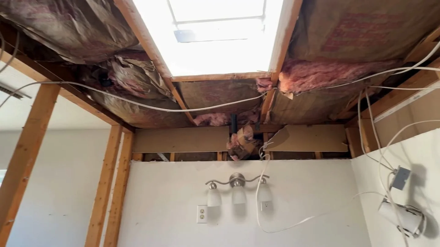 Opened ceiling above a bathroom showing wood framing, pink insulation, wiring and a skylight.