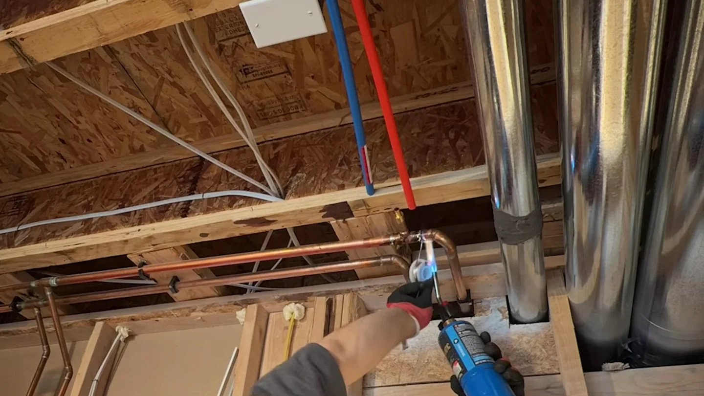 Opened ceiling with wood joists, red and blue PEX lines and a gloved hand holding a drill.