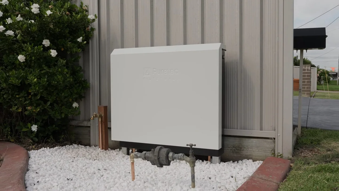White utility cabinet on a stucco wall beside a fence, with white gravel bed and pipes below.