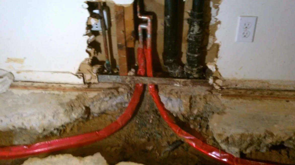 Two red PEX lines running down an opened wall to the slab beside dark drain pipes.