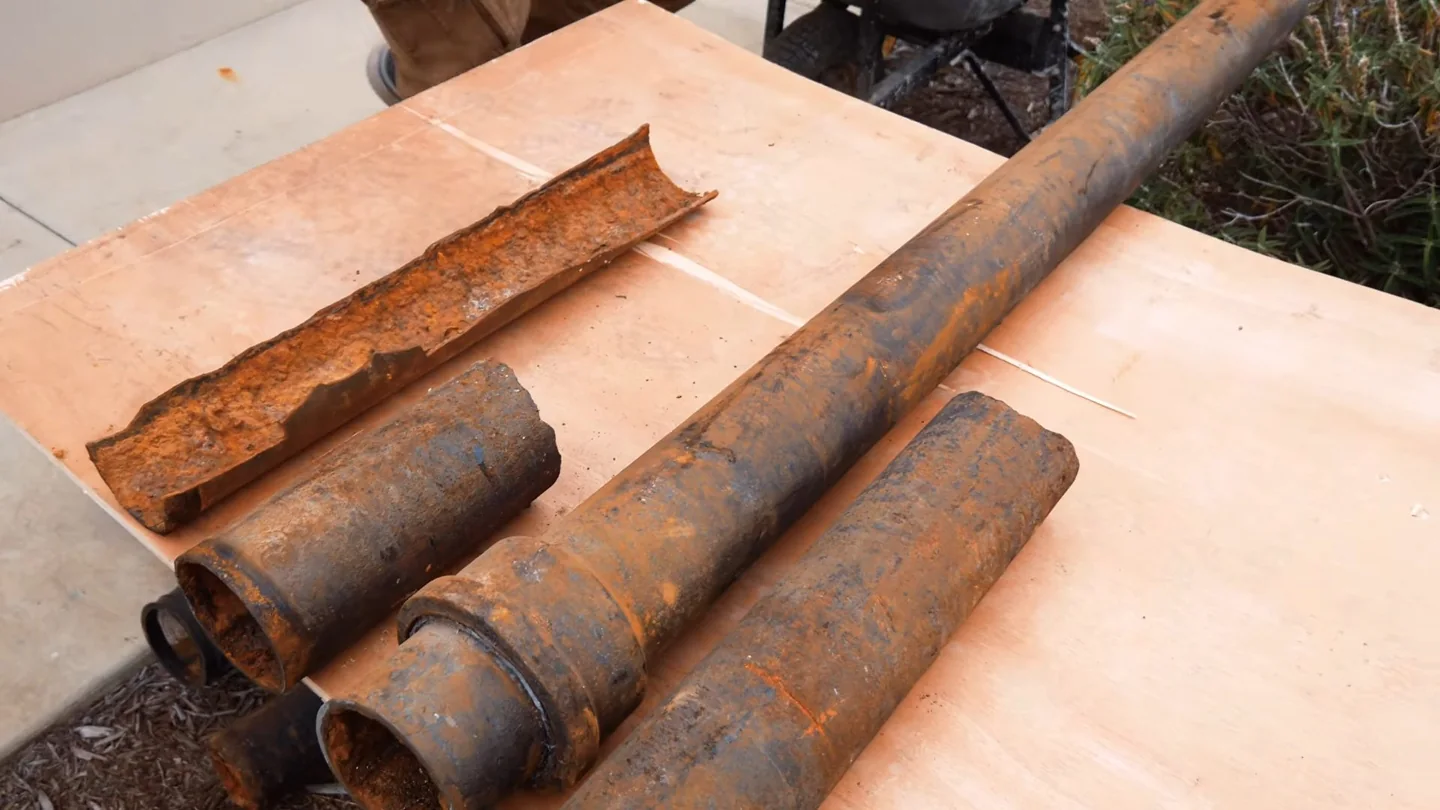 Rusted pipe sections, one split open lengthwise to show heavy corrosion inside, on a wooden table.