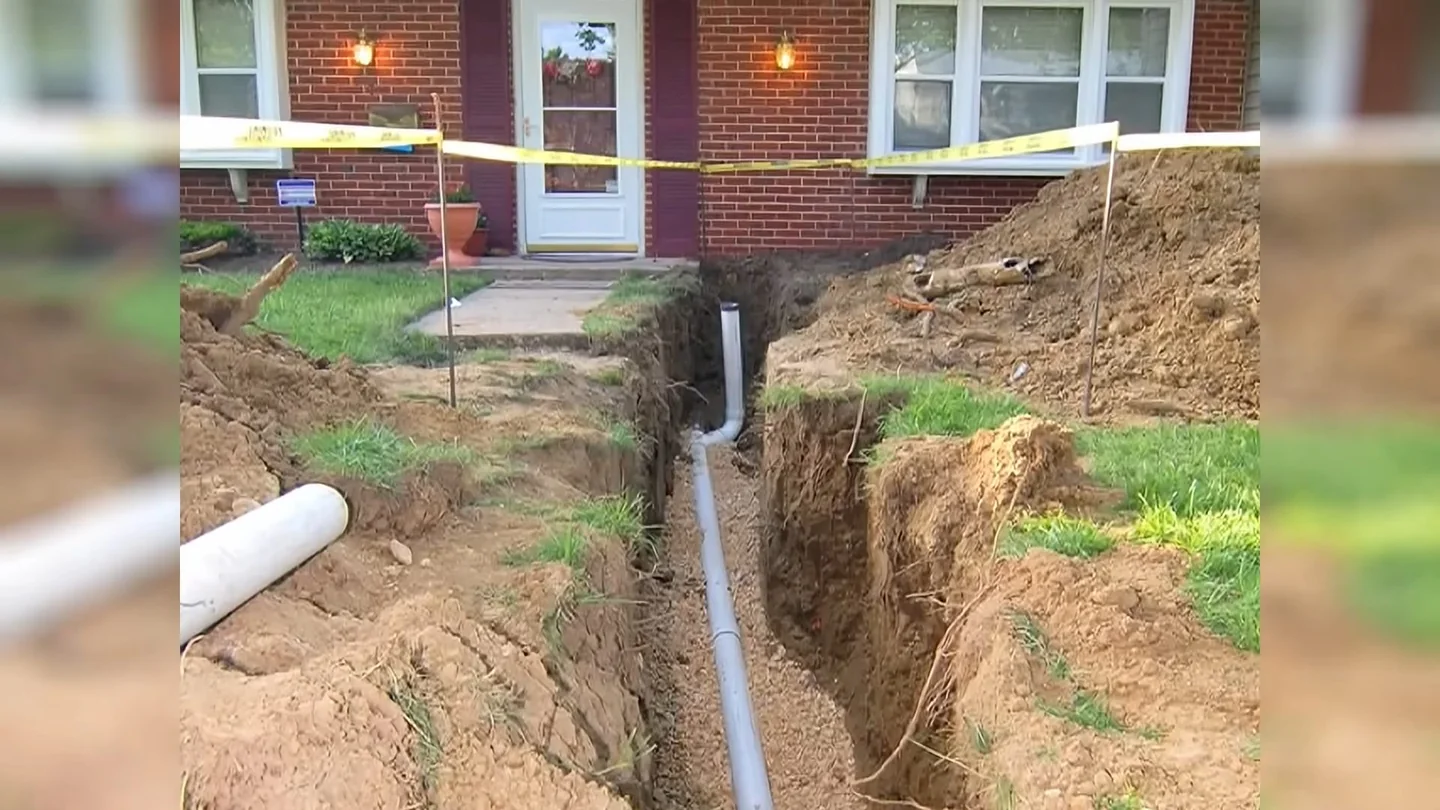 Trench with a plastic pipe running toward the front door of a brick house, with yellow tape along the yard.