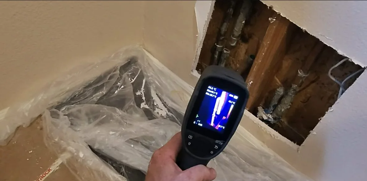 Hand holding a thermal camera toward an opened wall section where pipes are exposed.