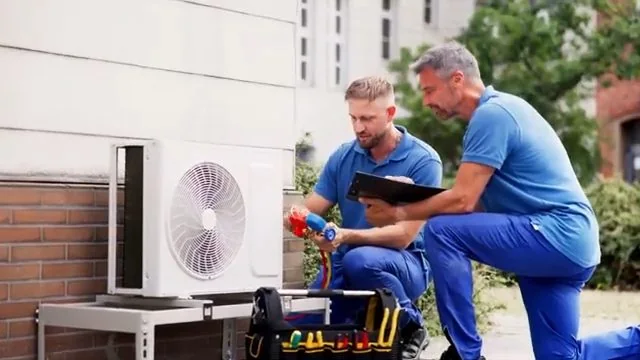 Two men in blue work shirts crouched beside an outdoor condenser unit, one holding a clipboard.