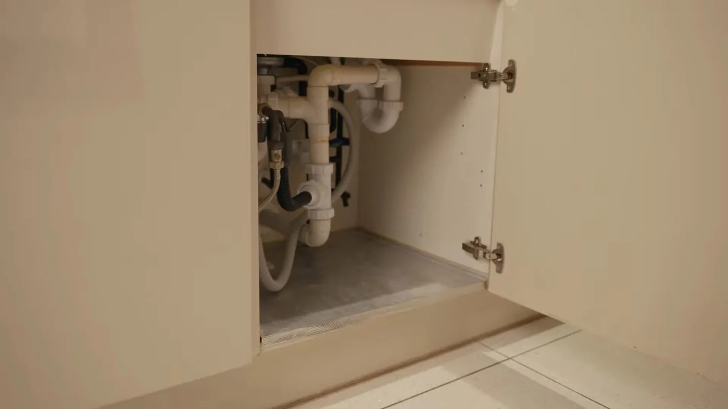 Open cabinet under a kitchen sink showing white drain pipes, a trap and supply lines.