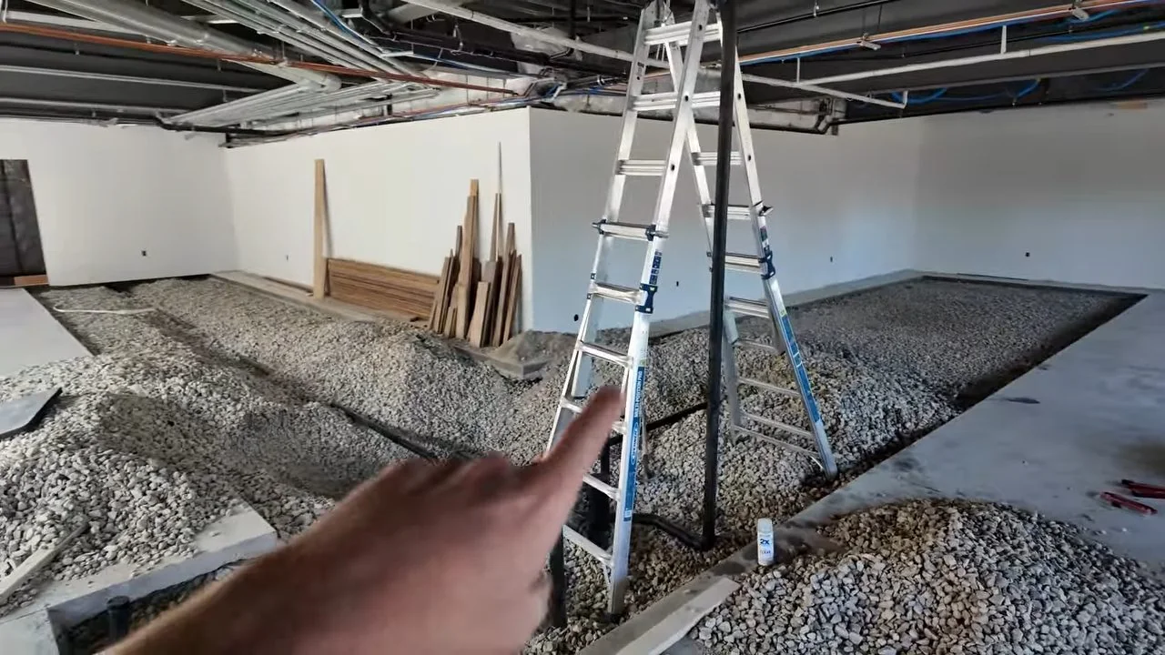 Unfinished commercial interior with a stepladder, a gravel-filled trench in the floor and pipes overhead.