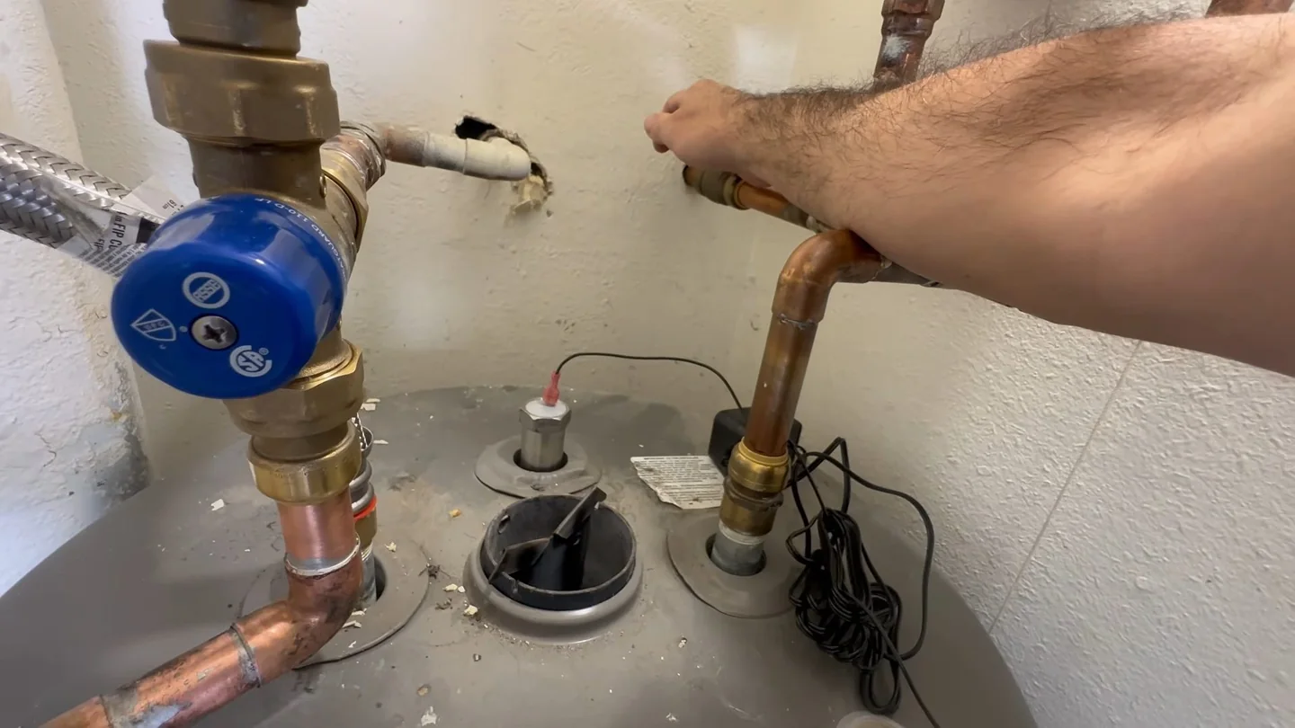 Hand reaching over a gray tank to copper lines, a blue valve and wiring on top of a water heater.