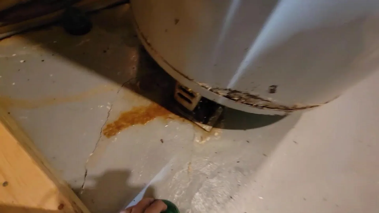 Base of a water heater with an orange rust stain on the floor and a hand reaching in.