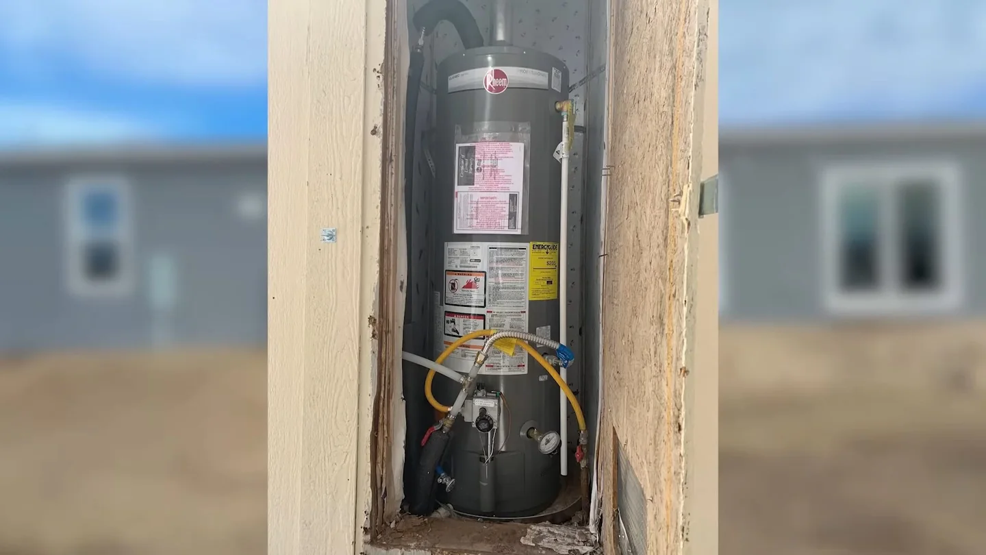 Water heater inside a framed wall opening, with a blurred row of modular homes in the background.