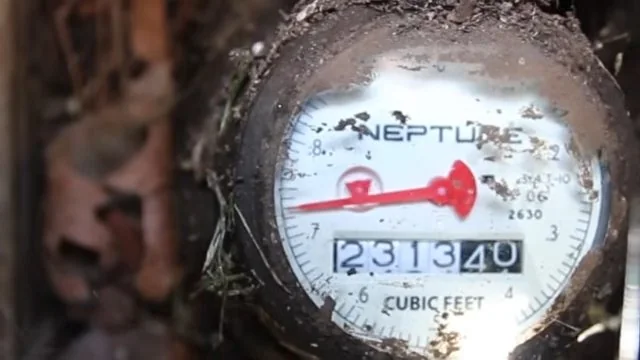 Water meter dial with a red sweep hand and a digit counter, seen inside a dirt-filled ground box.
